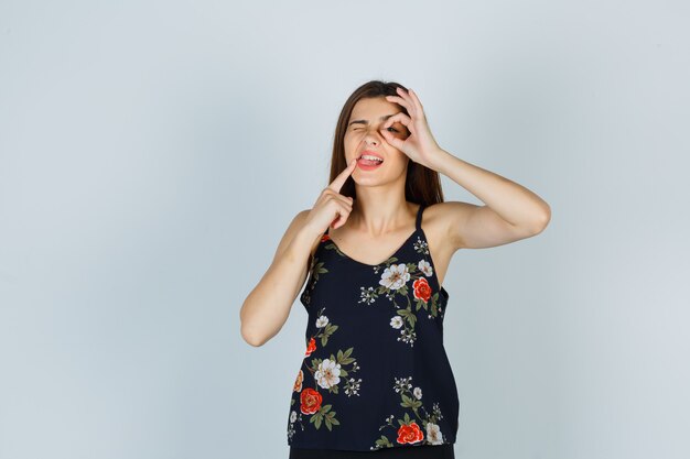 Attractive young lady showing ok sign on eye, holding finger near mouth, sticking out tongue while blinking in blouse and looking pensive. front view.