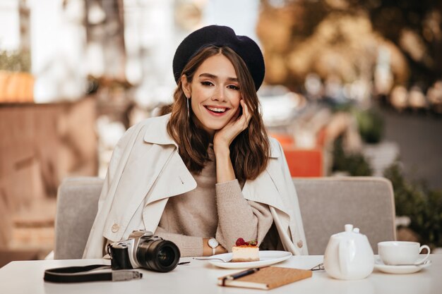 Attractive young lady with brunette wavy hairstyle, beret, beige trench coat having lunch at cafe terrace against sunny autumn city wall