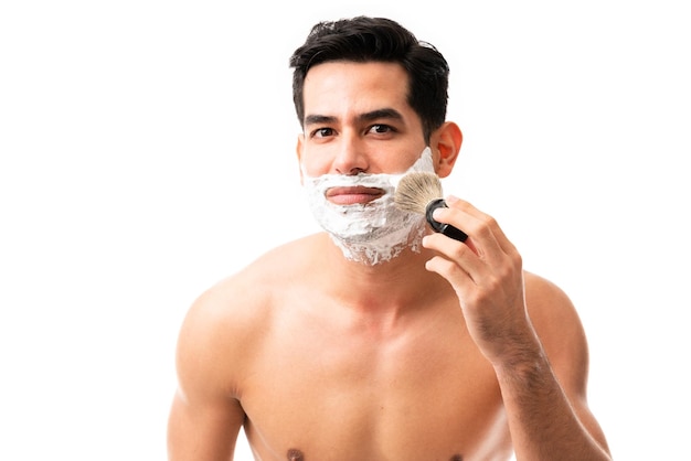 Привлекательный молодой мужчина наносит крем для бритья кистью на щеку на белом фоне