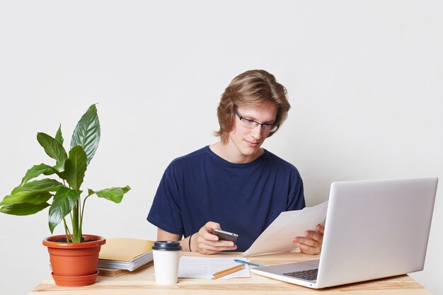 Attractive young male enterpreneur has serious look, textes message on mobile phone, uses free internet connection, does paper work during coffee break, isolated over white blank  wall