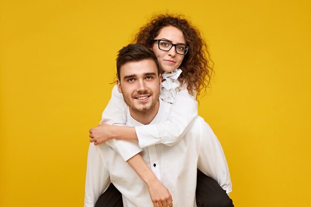 attractive young male with bristle giving piggyride to his adorable girlfriend in glasses. Stylish female riding on back of her boyfriend, smiling happily. Dating and relationships