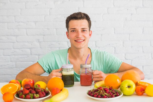 Attractive young man holding green and red smoothie jars with colorful fruits on table