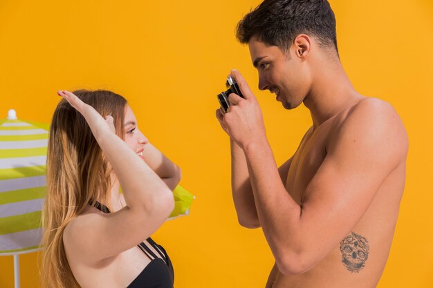 Attractive young man making photo of girlfriend in swimsuit on yellow background