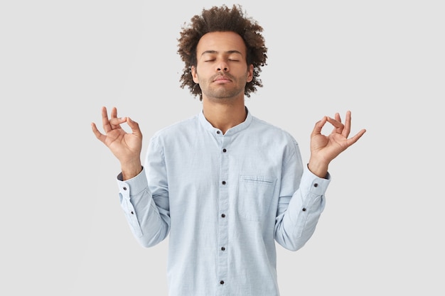 Attractive young man pracitices yoga, feels relaxed and calm, shows mudra sign with both hands, closes eyes as tries to concentrate on something, poses alone against white wall