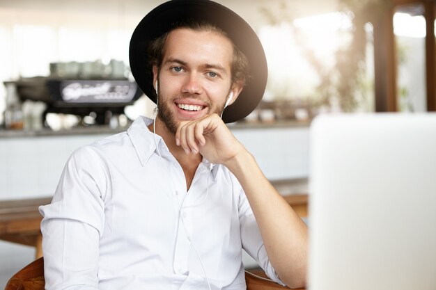 Привлекательный молодой человек расслабляется во время обеда в современном кафе, сидит перед открытым ноутбуком и счастливо улыбается, просматривая забавные видеоролики онлайн в наушниках