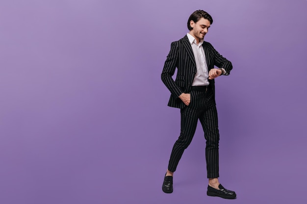 Привлекательный молодой человек в белой рубашке, черном полосатом костюме и модных туфлях улыбается и смотрит на часы на руке Образ жизни и бизнес-концепция на фиолетовом фоне