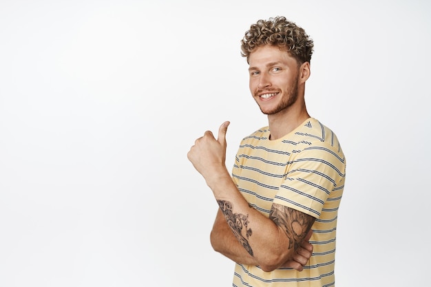 Attractive young man with beard and curly blond hairstyle shows thumbs up with proud satisfied face smiling pleased praising and complimenting say yes like smth white background
