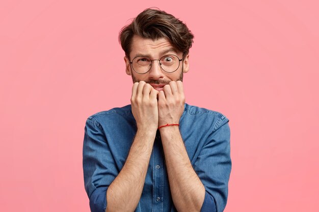 Attractive young man with confused nervous expression, bites finger nails, worries to make terrible mistake, feels anxious, looks embarrassed, wears denim shirt, isolated over pink wall