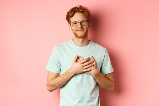 Attractive young man with ginger hair holding hands on heart and smiling grateful saying thank you e...
