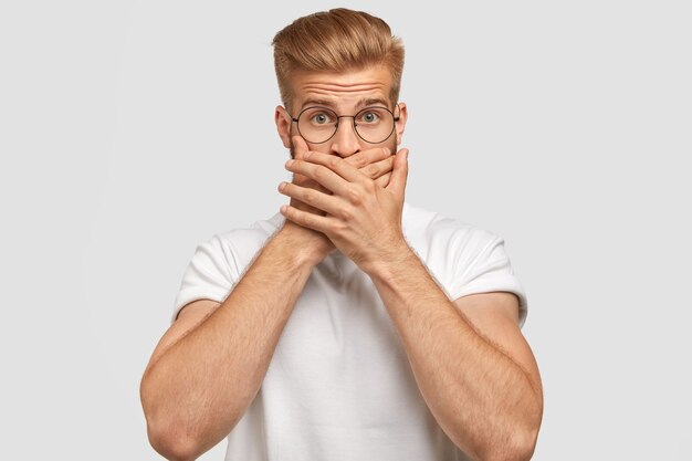 Attractive young man with trendy haircut, covers mouth, being mute