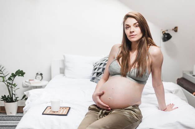 Attractive young pregnant woman sitting on bed touching her belly