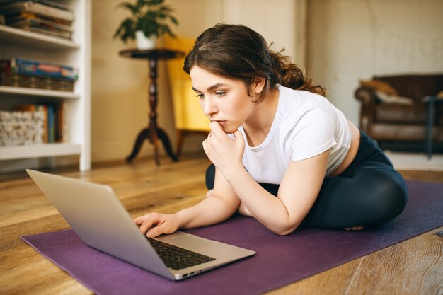 Attractive young professinal female fitness trainer sitting on mat with portable computer having pensive look, recording video class for online training. Pretty girl watching yoga tutorial on laptop