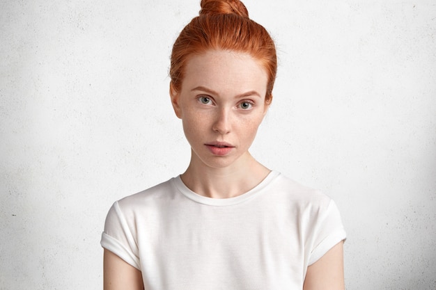 無料写真 そばかすのある肌と真剣な表情の魅力的な若い赤髪の女性。白いコンクリートの壁にカジュアルな白いtシャツを着たモデル。