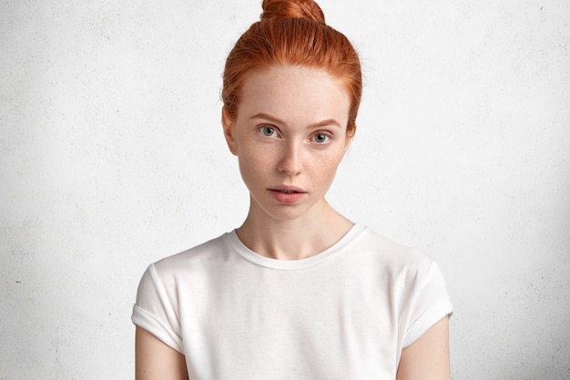 Attractive young red haired female with freckled skin and serious look, dressed in casual white t shirt, models against white concrete wall.