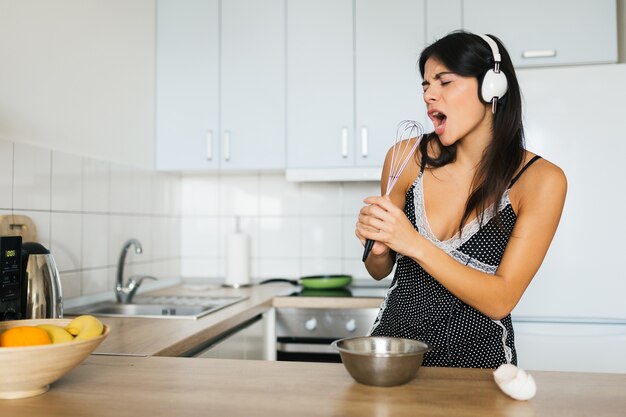 Attractive young skinny smiling woman having fun cooking eggs at kitchen in morning having breakfast dressed in pajamas outfit, listening to music on headphones singing