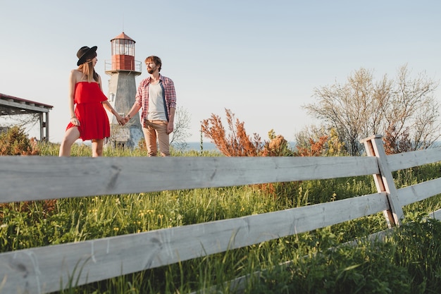 Free photo attractive young stylish couple in love in countryside, indie hipster bohemian style, weekend vacation, summer outfit, red dress, green grass, holding hands