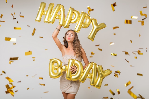 Attraente giovane donna elegante che celebra, che tiene le lettere di buon compleanno degli aerostati di aria, coriandoli dorati che volano, sorridendo felice, isolato, vestito da festa da portare