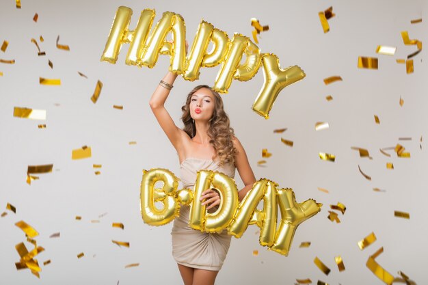 Attractive young stylish woman celebrating, holding air balloons happy birthday letters, golden confetti flying, smiling happy, isolated, wearing party dress
