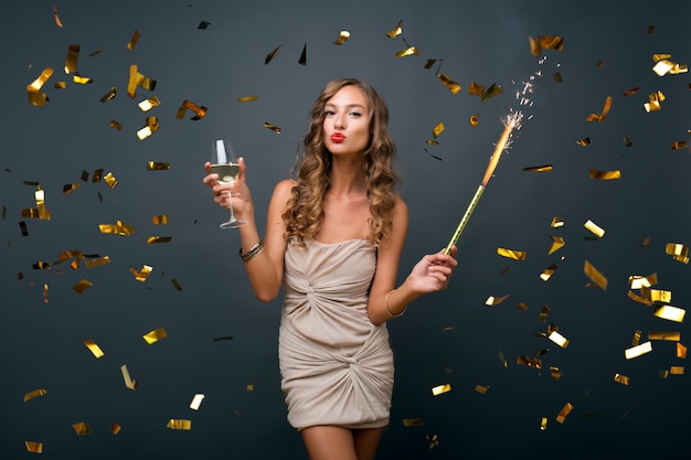 Attraente giovane donna elegante che celebra il nuovo anno, bevendo champagne, coriandoli dorati che volano, sorridendo felice, isolato, con indosso un abito da festa