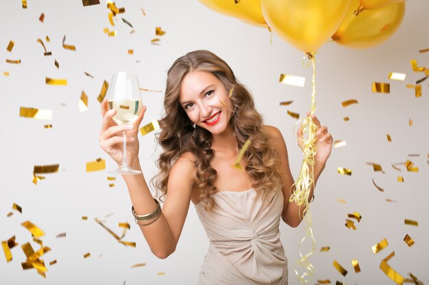Attractive young stylish woman celebrating new year, drinking champagne holding air balloons, golden confetti flying, smiling happy, isolated, wearing party dress