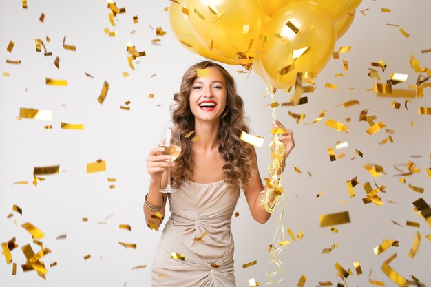 Attractive young stylish woman celebrating new year, drinking champagne holding air balloons, golden confetti flying, smiling happy, white, isolated, wearing party dress