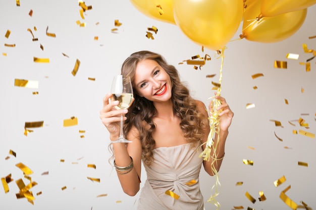 Attractive young stylish woman celebrating new year, drinking champagne holding air balloons, golden confetti flying, smiling happy, white, isolated, wearing party dress