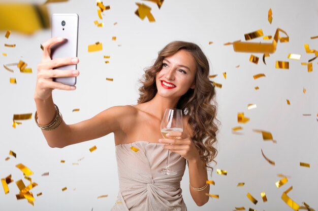 Attractive young stylish woman celebrating new year, drinking champagne making selfie photo on phone, golden confetti flying, smiling happy, isolated, wearing party dress