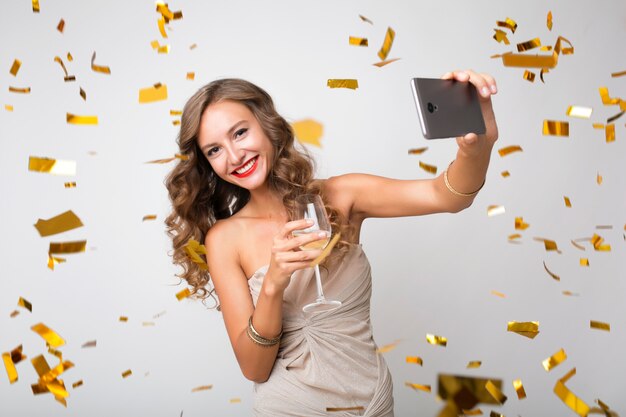 魅力的な若いスタイリッシュな女性が新年を祝って、電話でシャンパンを作るselfie写真を飲んで、金色の紙吹雪が飛んで、笑って幸せ、分離、パーティードレスを着て