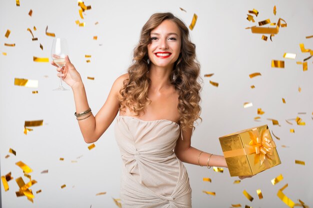 Привлекательная молодая стильная женщина празднует новый год, держит подарки в коробке, летит золотое конфетти, улыбается счастливой, в вечернем платье