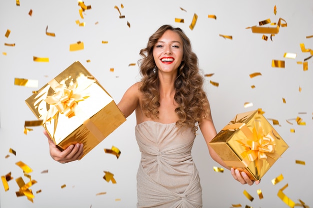 Attractive young stylish woman celebrating new year, holding presents in box, golden confetti flying, smiling happy, wearing party dress