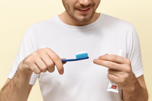 Attractive young unshaven male posing isolated holding toothbrush with brushing gel in it, going to do morning routine, smiling at camera, showing teeth. Cropped image