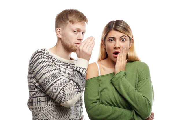 Attractive young unshaven male sharing secret or whispering gossips into his astonished girlfriend's ear, who is staring with mouth wide opened, shocked with unexpected information
