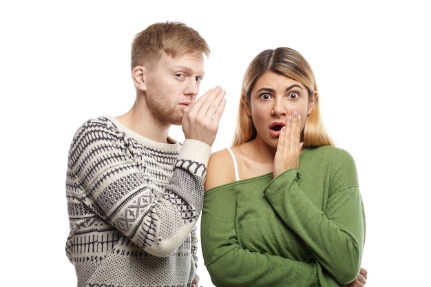 Attractive young unshaven male sharing secret or whispering gossips into his astonished girlfriend's ear, who is staring with mouth wide opened, shocked with unexpected information