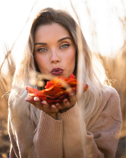 Free photo attractive young woman blowing autumn leaves at outdoors looking at camera