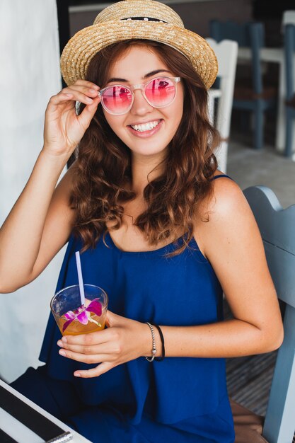 Attractive young woman in blue dress and straw hat wearing pink sunglasses, drinking alcohol cocktail on tropical vacation and sitting at table in bar