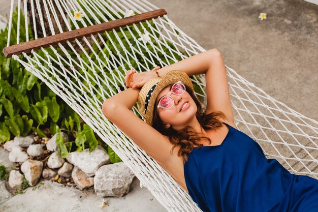 파란 드레스와 해먹에 누워 휴가에 편안한 핑크 선글라스를 쓰고 밀짚 모자에 매력적인 젊은 여자