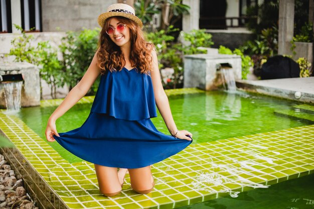 Foto gratuita attraente giovane donna in abito blu e cappello di paglia che indossa occhiali da sole rosa in piscina