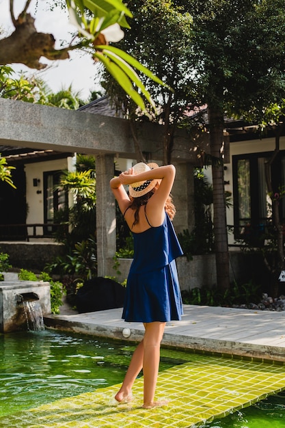 Free photo attractive young woman in blue dress and straw hat wearing pink sunglassses walking at pool of tropical spa villa on vacation in summer style outfit, view from back holding hands up