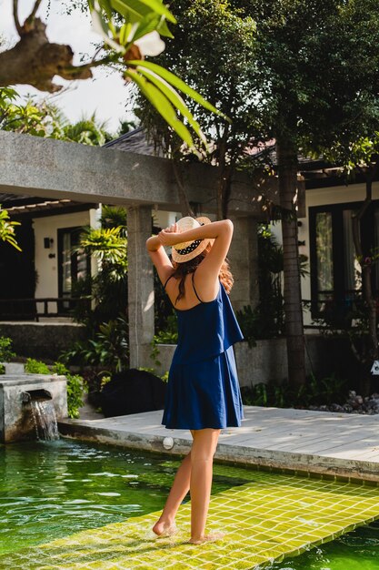 Attractive young woman in blue dress and straw hat wearing pink sunglassses walking at pool of tropical spa villa on vacation in summer style outfit, view from back holding hands up