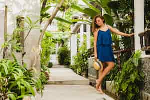 Free photo attractive young woman in blue dress and straw hat wearing pink sunglassses walking at tropical spa villa hotel on vacation in summer style outfit, sexy