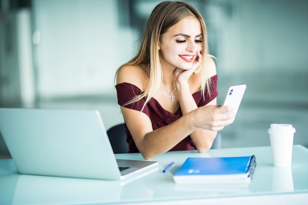 Привлекательная молодая женщина с серьезным выражением лица проверяет свои текстовые сообщения на мобильном телефоне, сидя за своим столом в офисе