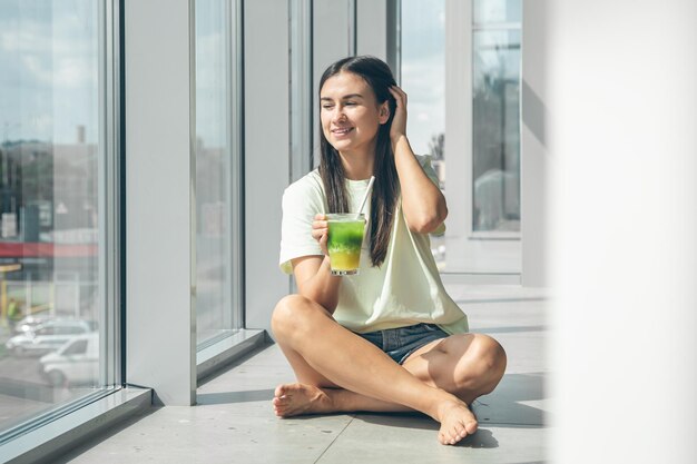 Free photo attractive young woman drinking a cooling drink while sitting by the window