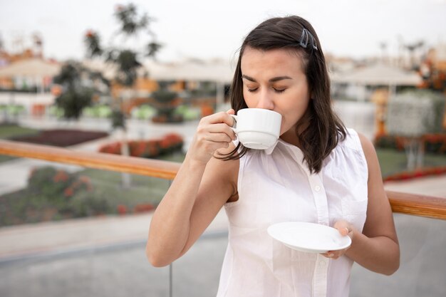 一杯のコーヒーと受け皿を手に屋外で朝を楽しんでいる魅力的な若い女性。休暇とレクリエーションの概念。