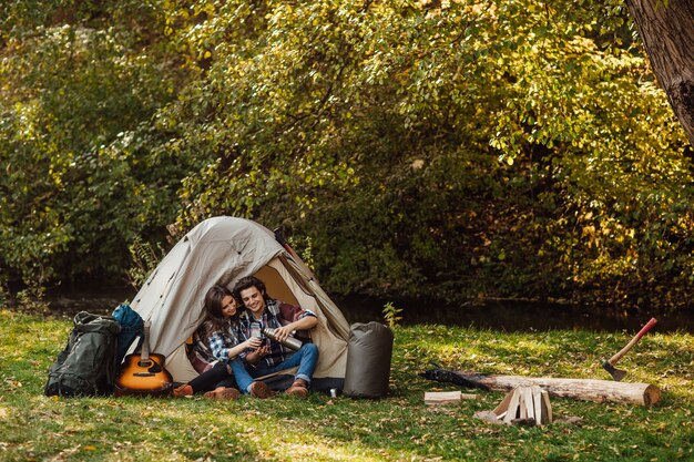 Attractive young woman and handsome man are spending time together on nature. Sitting in touristic tent in forest and drinking tea