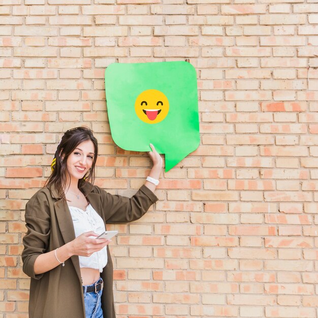 Attractive young woman holding cellphone and happy emoji message against brick wall