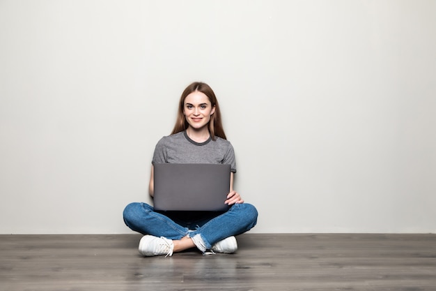 Attractive young woman holding laptop computer while sitting on the floor with legs crossed and looking away at copy space isolated over gray wall