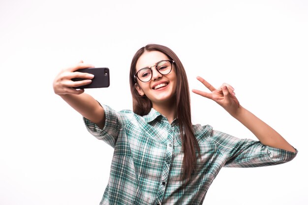 Attractive young woman holding mobile phone and making photo of herself while standing against white