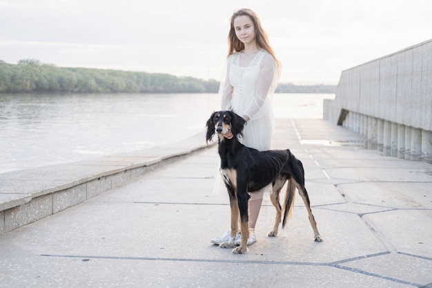白いドレスの魅力的な若い女性は、街でサルーキ犬と一緒に歩きます。川岸の近く。ペルシャグレイハウンド。ペットケアのコンセプト。人間と動物の間の愛と友情。 Premium写真