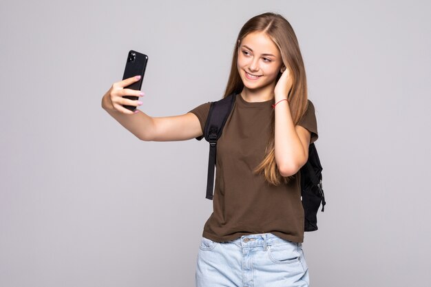 Attractive young woman making selfies on mobile phone camera on white wall