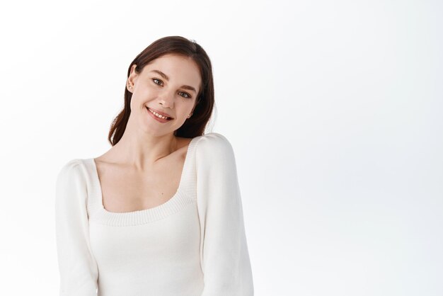 Attractive young woman posing cute on white studio backround tilt head and smile at camera softly making gentle face expression standing over white background